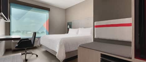 A hotel room with a bed, a desk, a chair, a mini-fridge, and a large window offering a view.