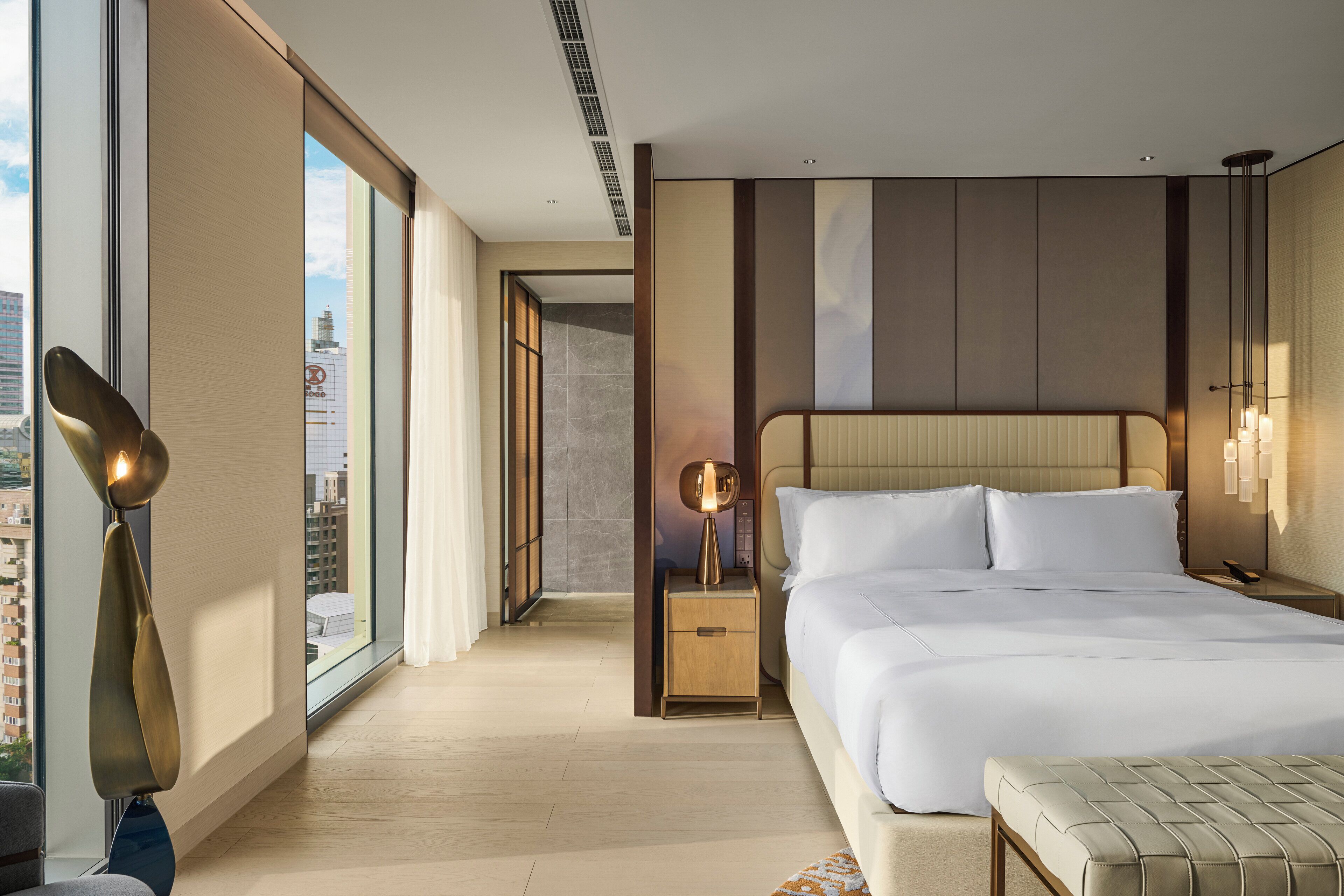 A modern hotel room with a large bed, bedside tables, a lamp, and a view of the city.