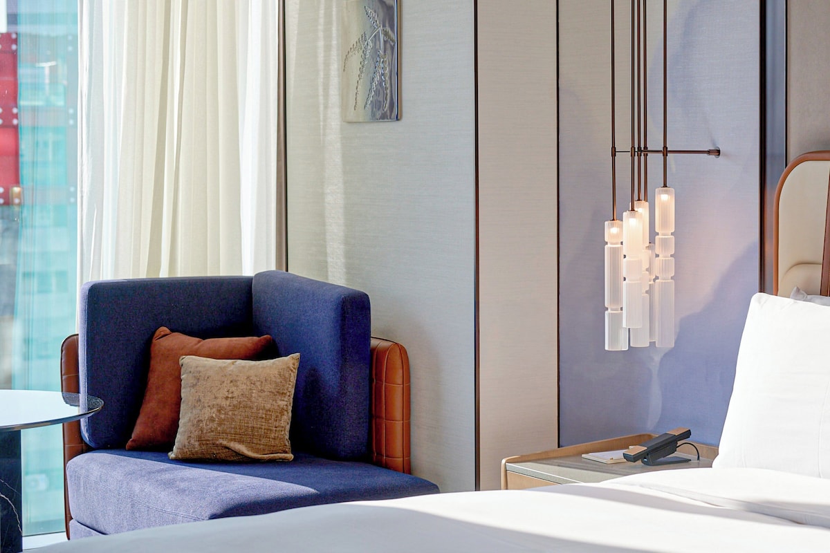 Une chambre d’hôtel avec un lit, un fauteuil bleu avec un coussin marron, une table de chevet avec une lampe et un luminaire fixé au mur.
