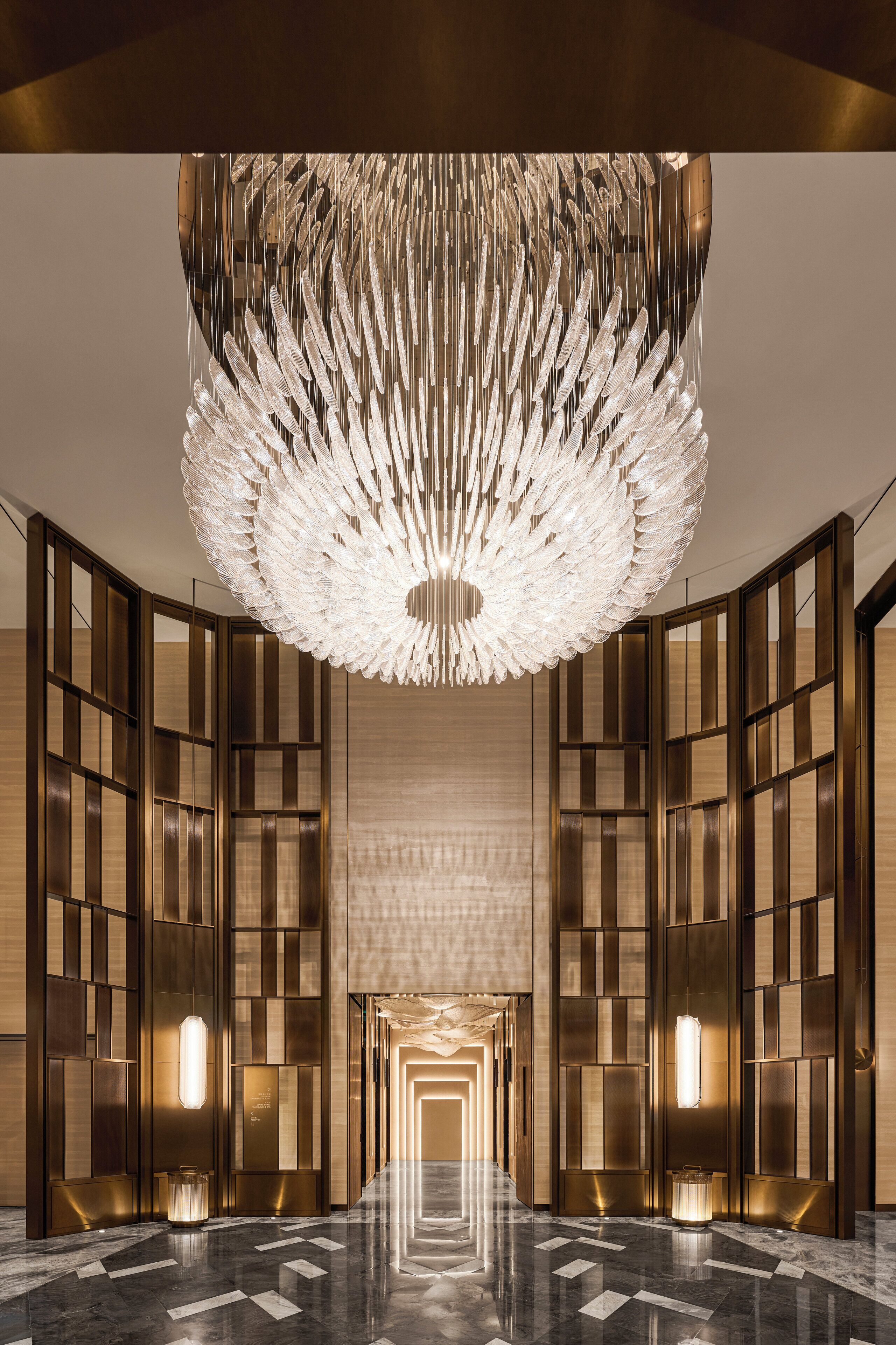 A modern hotel lobby with a central chandelier, mirrored walls, and a geometric floor pattern.