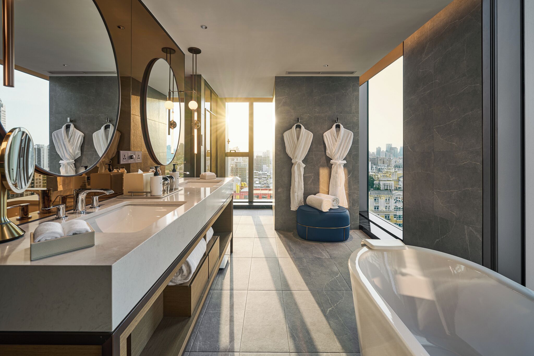 A modern bathroom with a large mirror, dual sinks, a bathtub, and a city view.