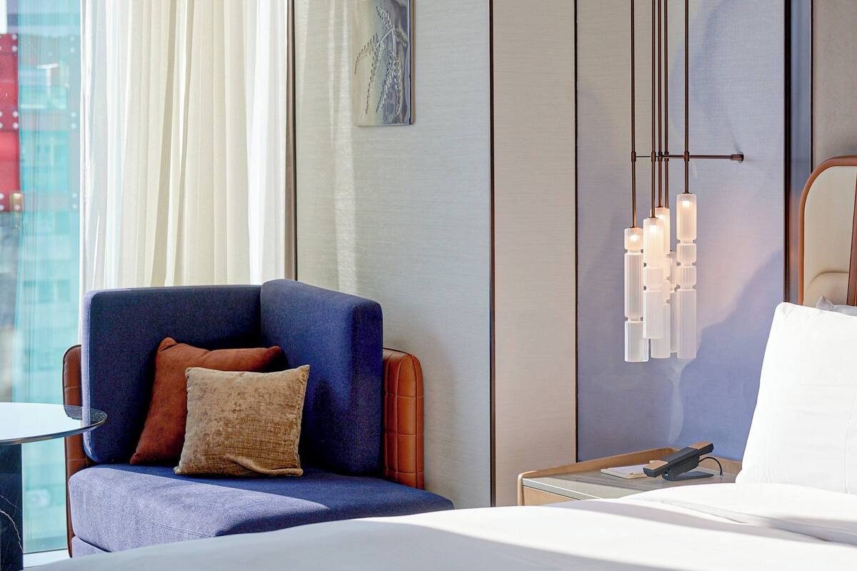 Una habitación de hotel con una cama, un sillón azul con un cojín marrón, una mesita de noche con una lámpara y una lámpara de pared.