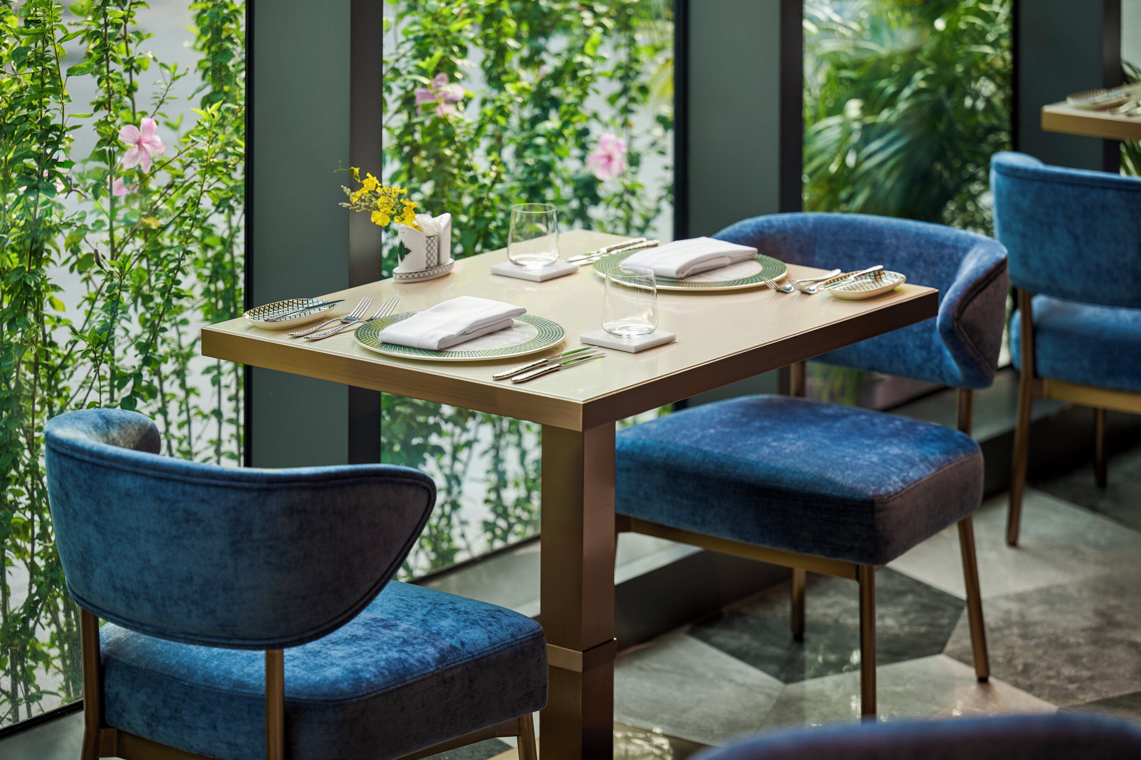 A dining area with a table set for two, blue upholstered chairs, and a view of greenery through a window.