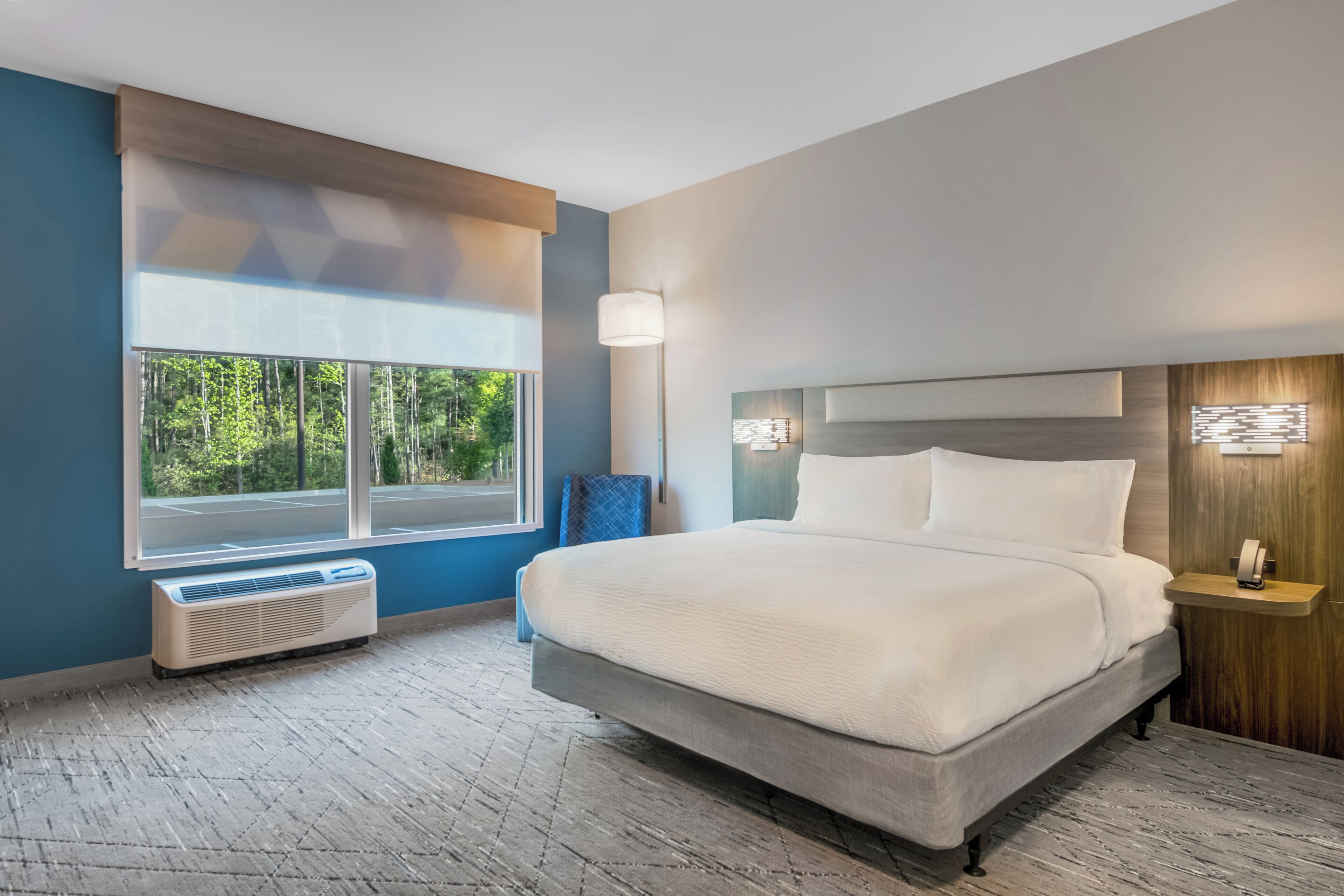 Habitación de hotel moderna con una cama grande, una mesita de noche, una lámpara y vistas al exterior.