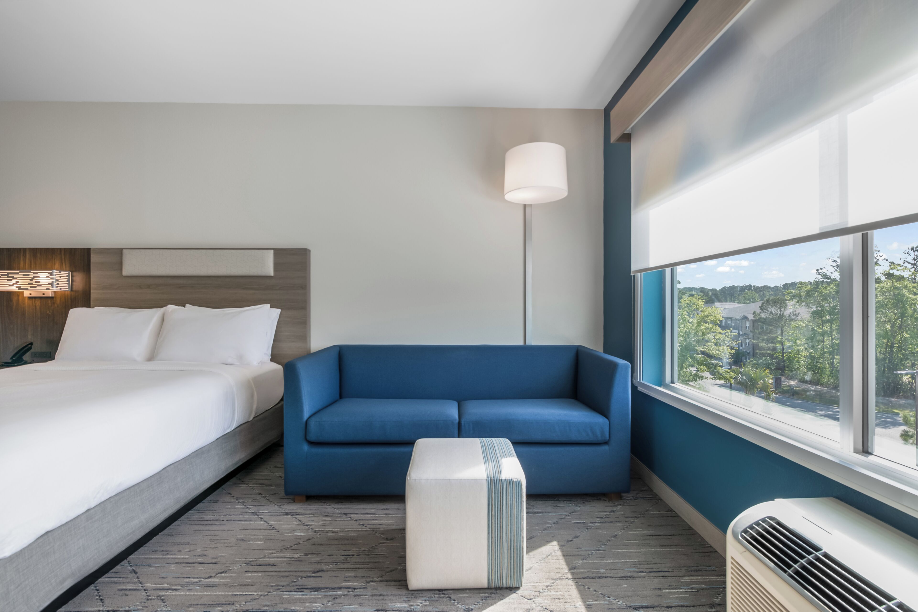Une chambre d’hôtel avec un lit, un canapé bleu, une lampe sur pied, une fenêtre donnant sur un paysage agréable et un radiateur.