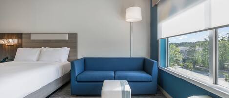 Une chambre d’hôtel avec un lit, un canapé bleu, une lampe sur pied, une fenêtre donnant sur un paysage agréable et un radiateur.