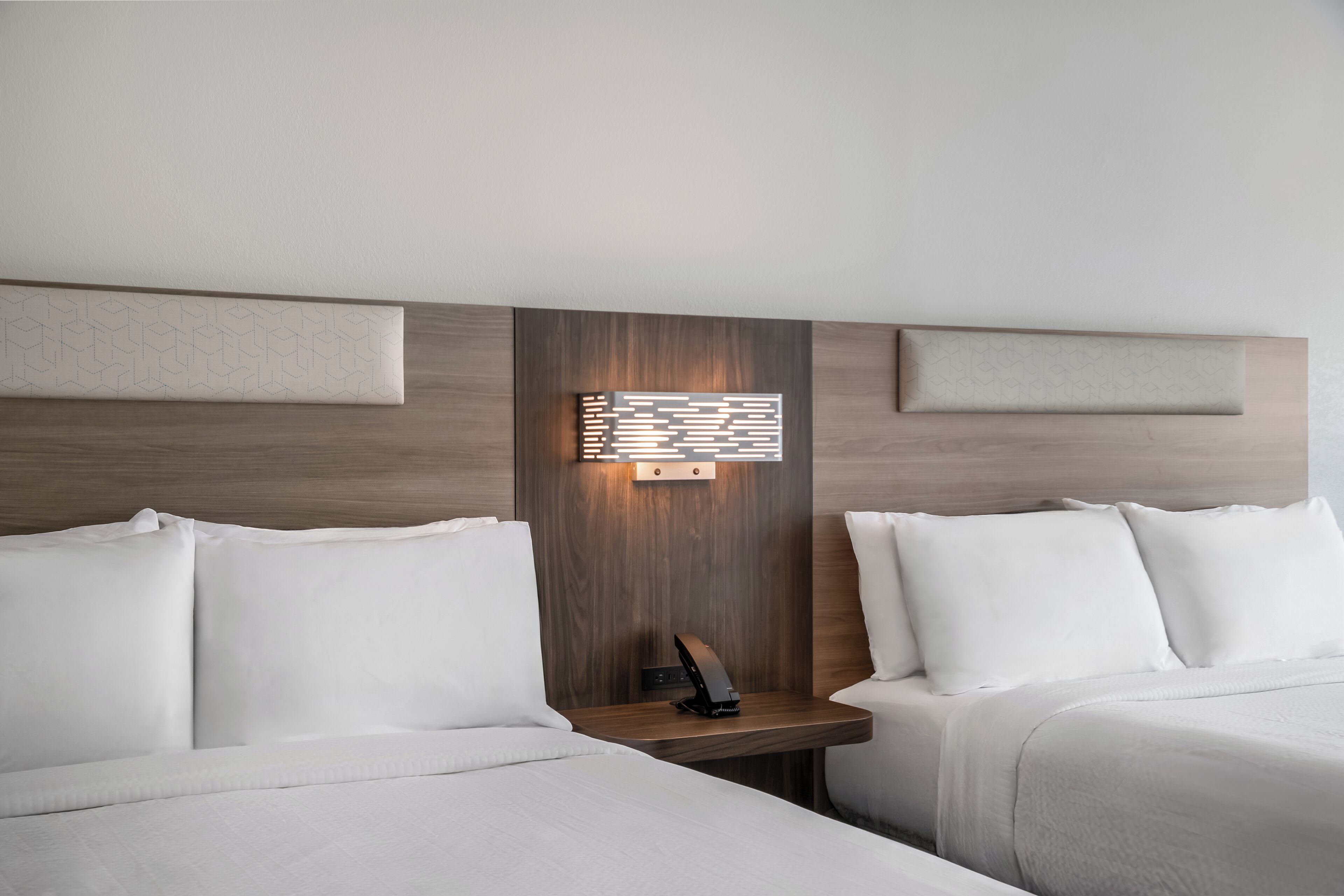 A hotel room with two beds, a bedside table with a lamp, and a wall-mounted light fixture.