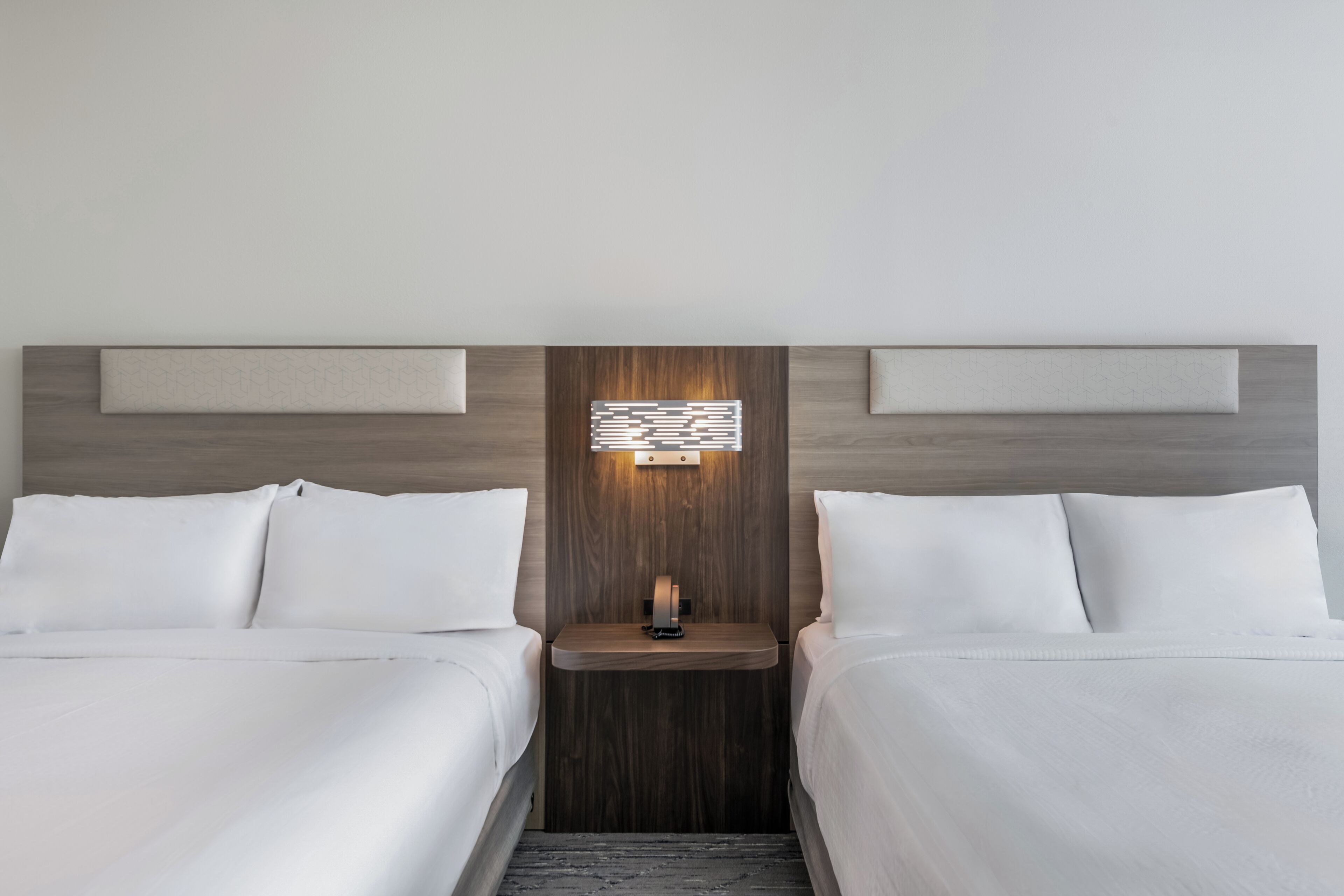 A hotel room with two beds, a nightstand, a wall-mounted light, and a wooden headboard.