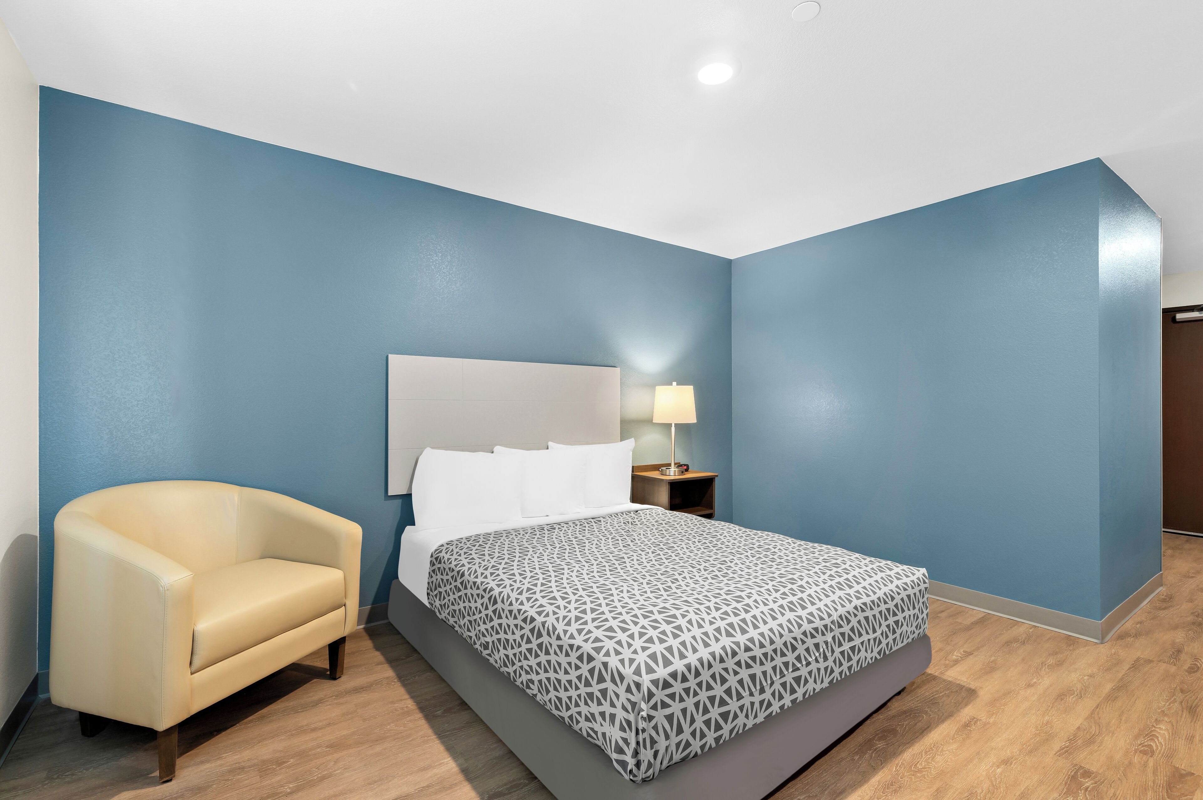 Una habitación de hotel con cama, una silla, una mesita de noche con lámpara y paredes azules.