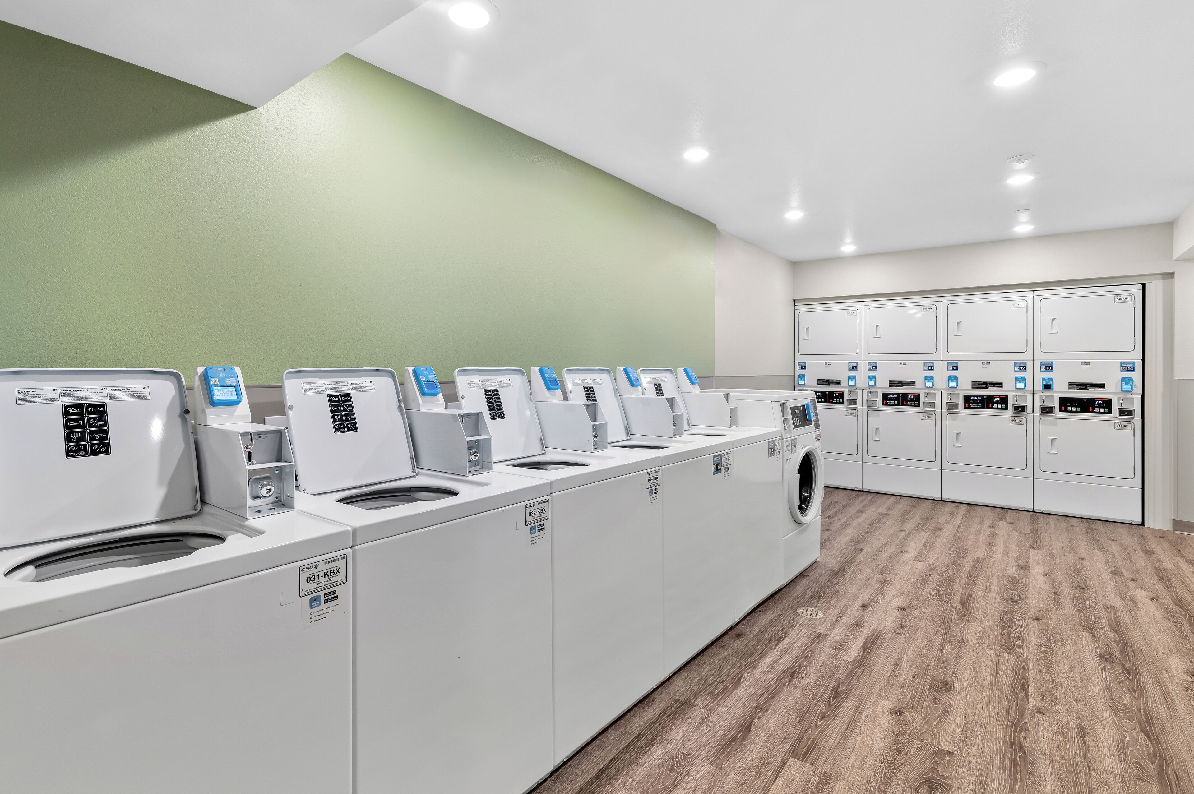 Une rangée de machines à laver et de sèche-linge modernes à chargement frontal, de couleur blanche, dans une buanderie bien éclairée, avec un mur d’accent vert et un plancher en bois.