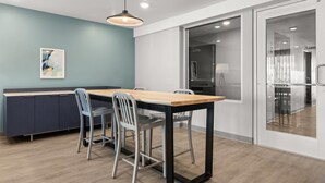 A modern kitchen with a wooden table and chairs, a blue accent wall, and a glass door leading to another room. - Woodspring Suites Ceres - Modesto (Ceres)