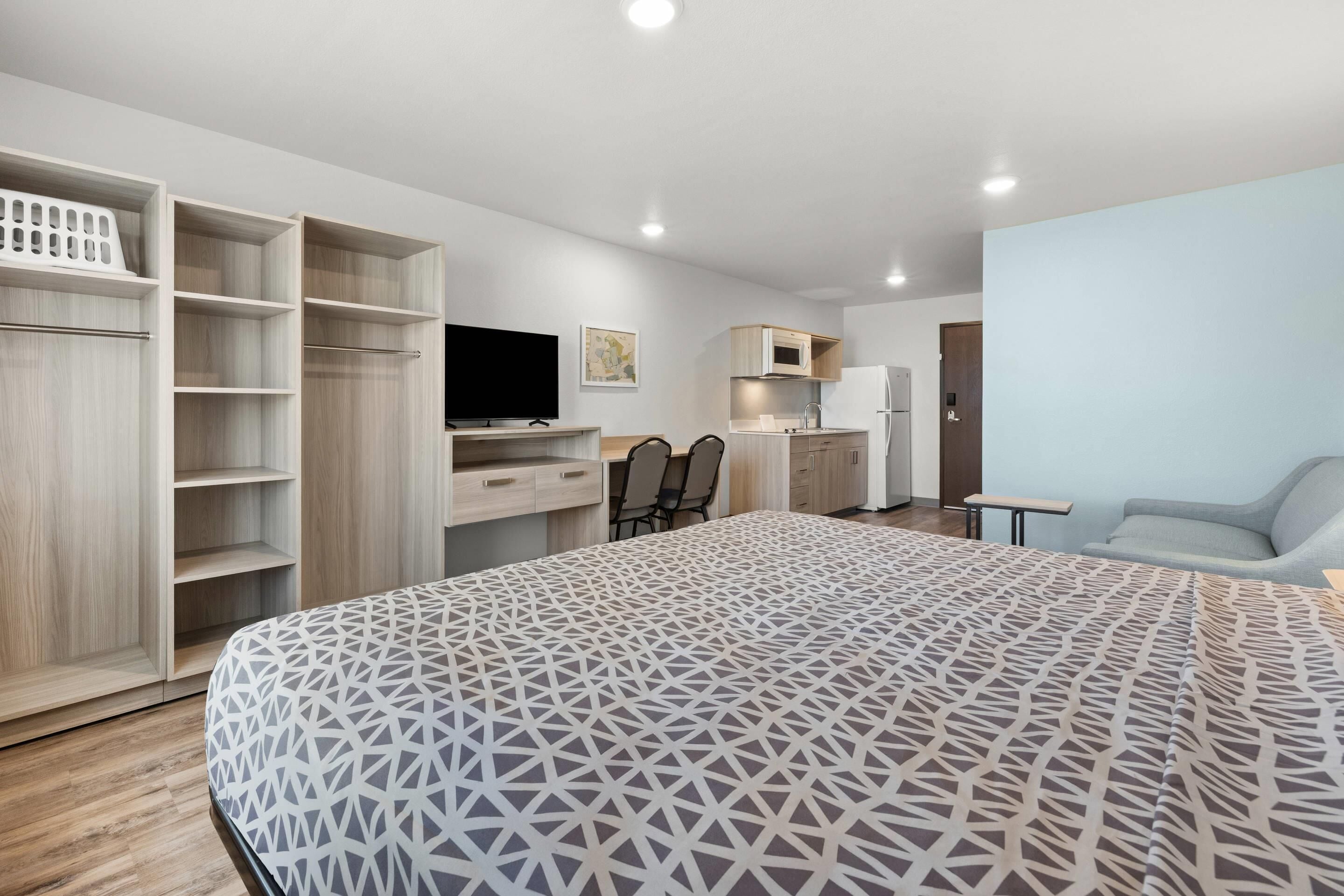 A modern bedroom with a large bed, a TV, a desk, and a kitchenette.
