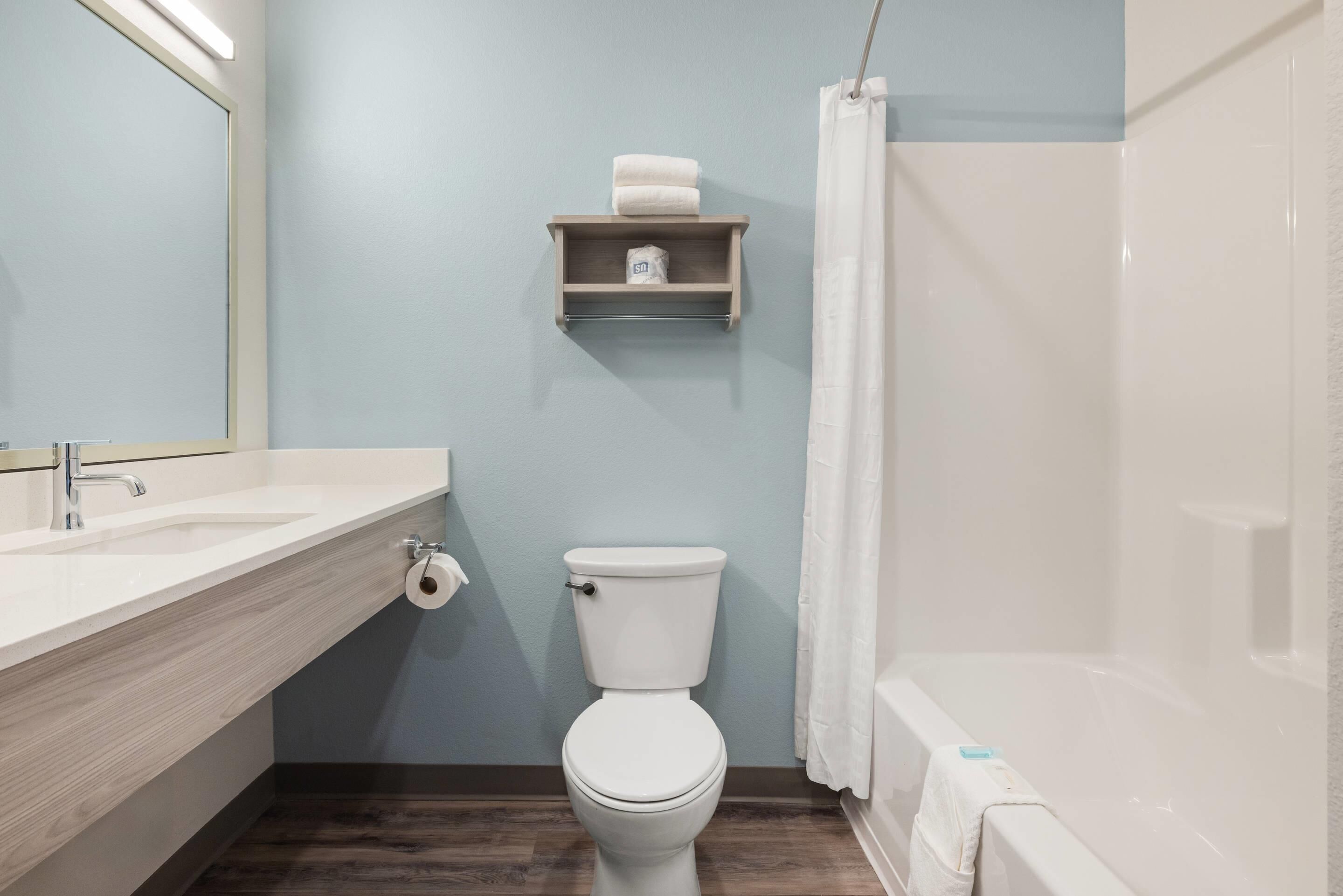 A bathroom with a white toilet, a wooden vanity with a mirror, a shelf with a towel and toilet paper, and a shower curtain.