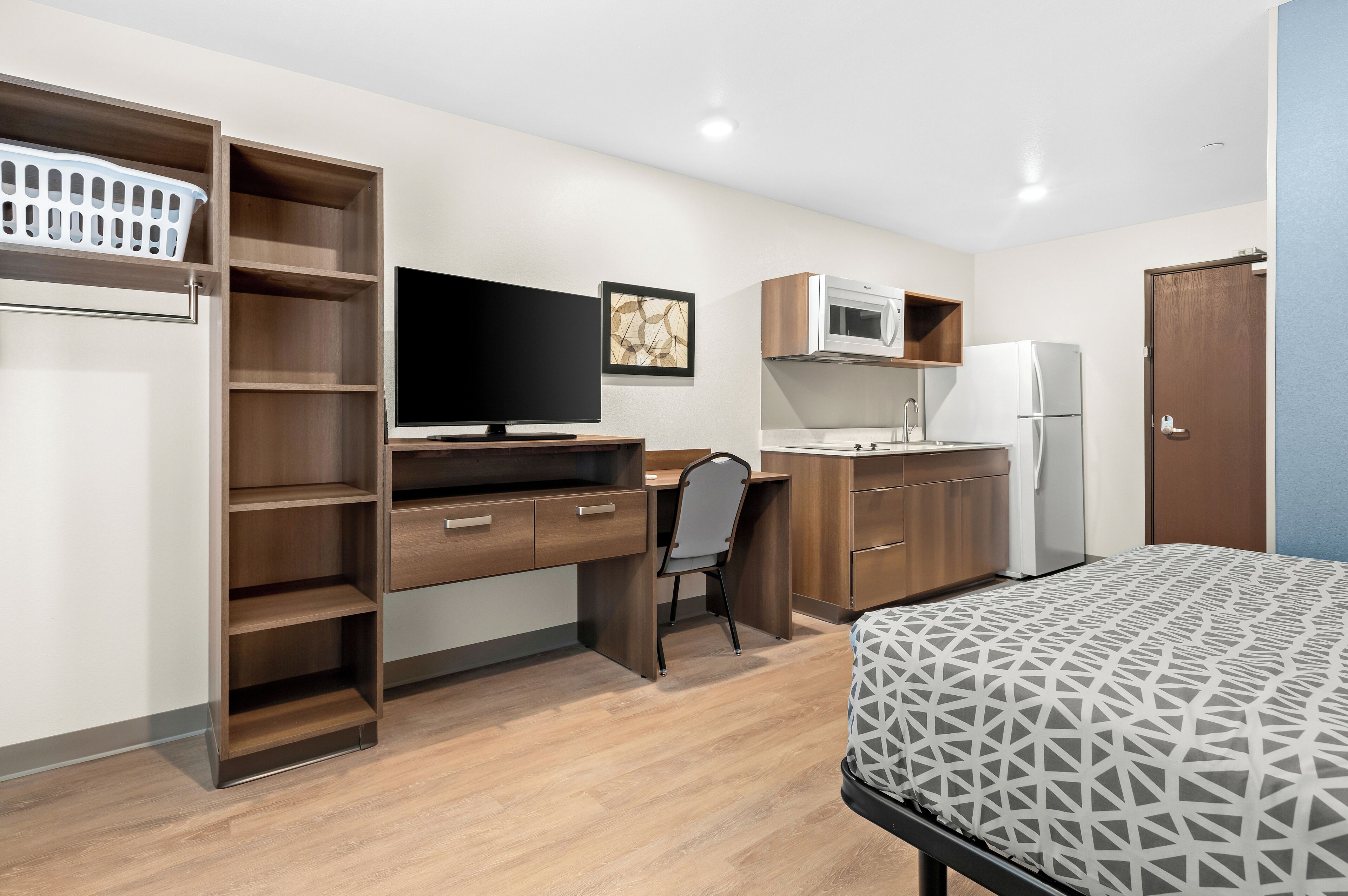 Une pièce de taille modeste comprenant un lit, un bureau avec une chaise, une télévision, un four à micro-ondes, un réfrigérateur et un panier à linge.