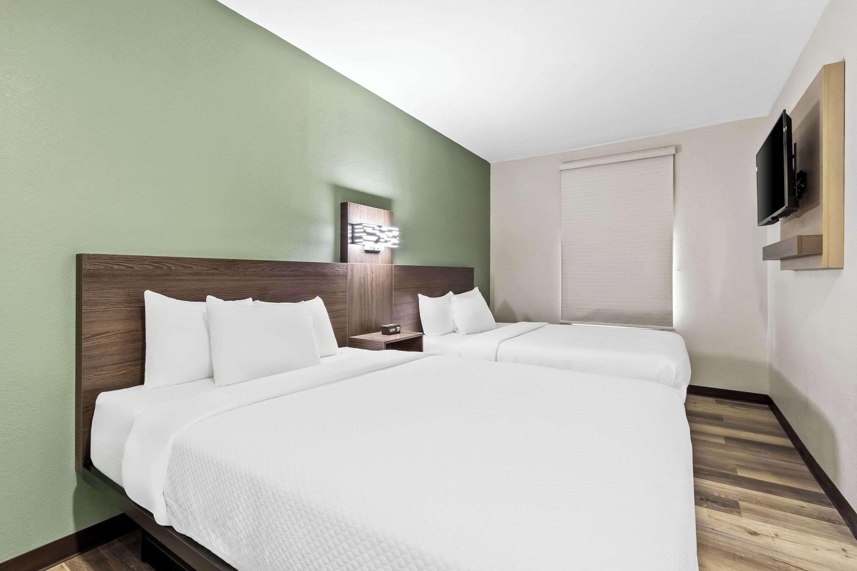 Habitación de hotel con dos camas, cabecero de madera, mesita de noche con lámpara, televisor de pantalla plana montado en la pared y ventana con persianas.