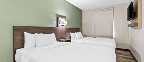 Habitación de hotel con dos camas, cabecero de madera, mesita de noche con lámpara, televisor de pantalla plana montado en la pared y ventana con persianas.