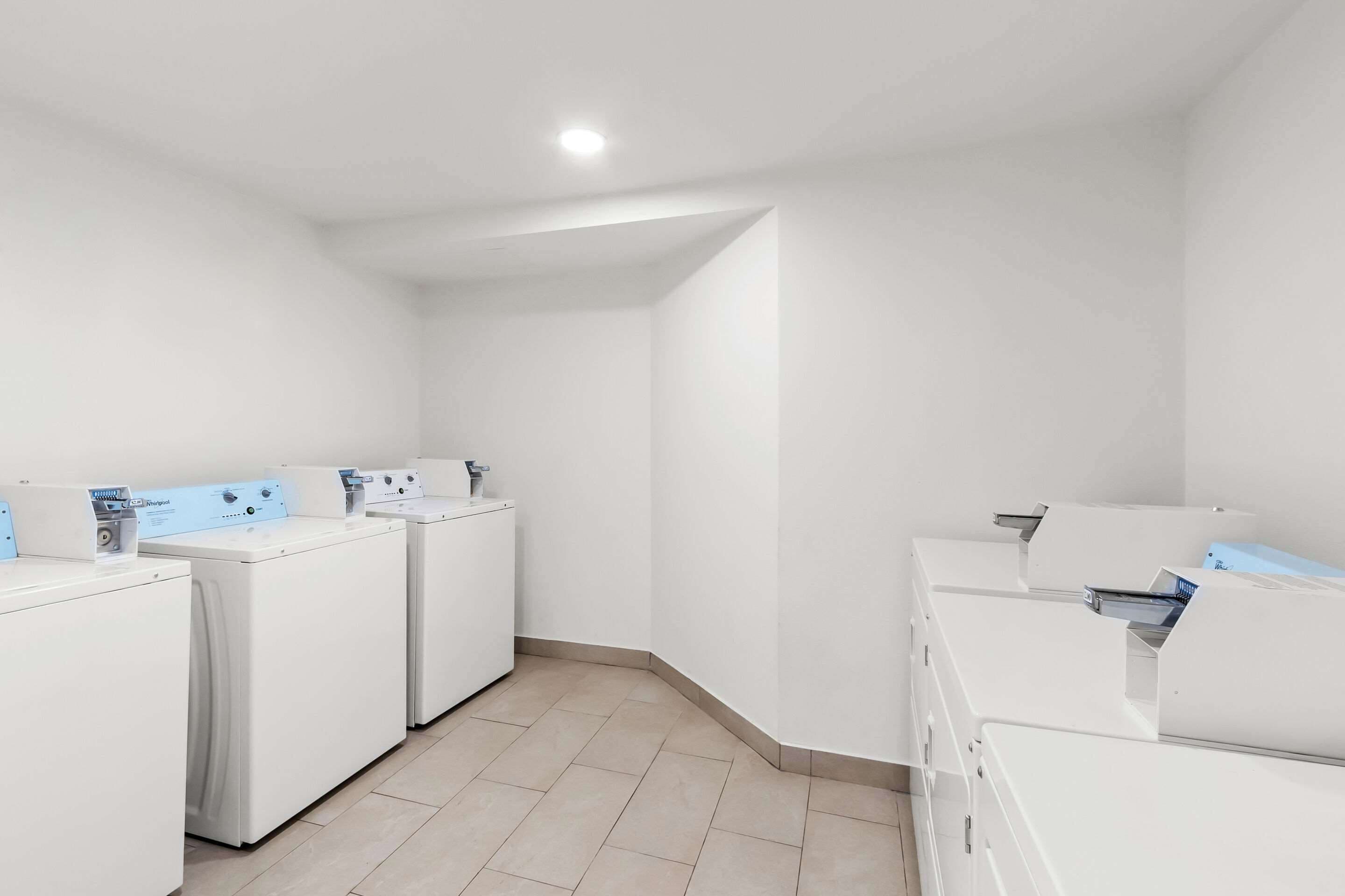 Une buanderie équipée de quatre machines à laver et de quatre sèche-linge blancs, avec un sol carrelé et un luminaire encastré au plafond.