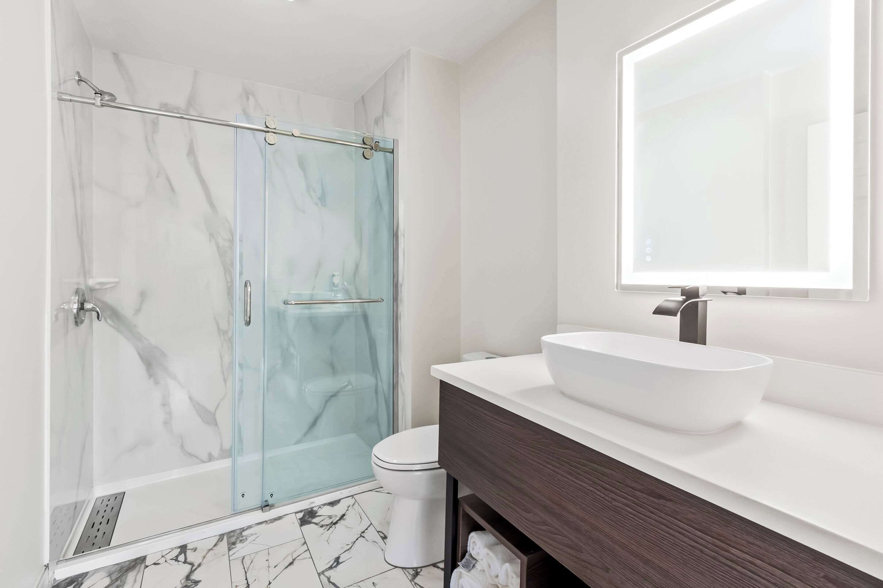 A modern bathroom with a glass-enclosed shower, a white sink with a large mirror, and a marble-tiled floor.