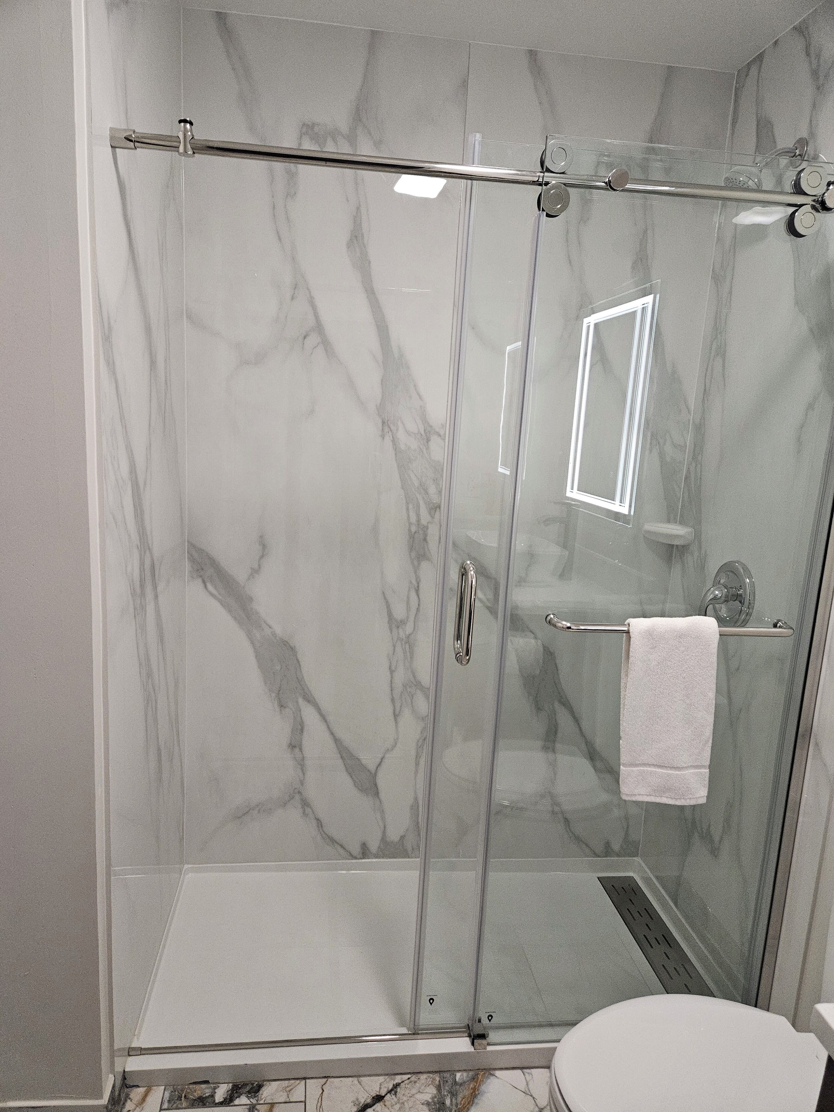 Ducha envuelta en vidrio con pared de mármol, puerta de cristal y una toalla blanca colgada en un perchero.