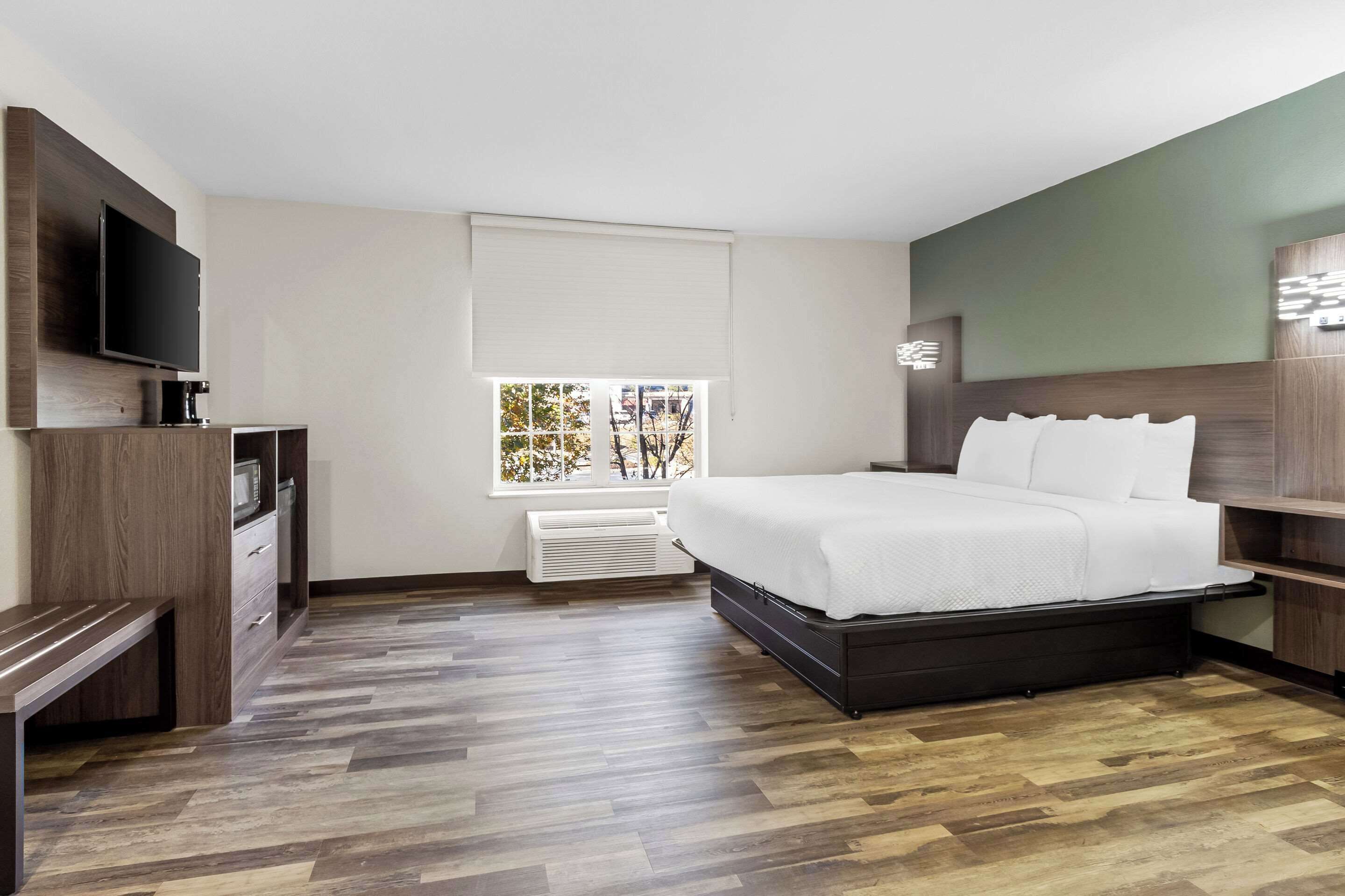 A modern hotel room with a large bed, a flat-screen TV, a built-in bench, and a window with a view of trees.