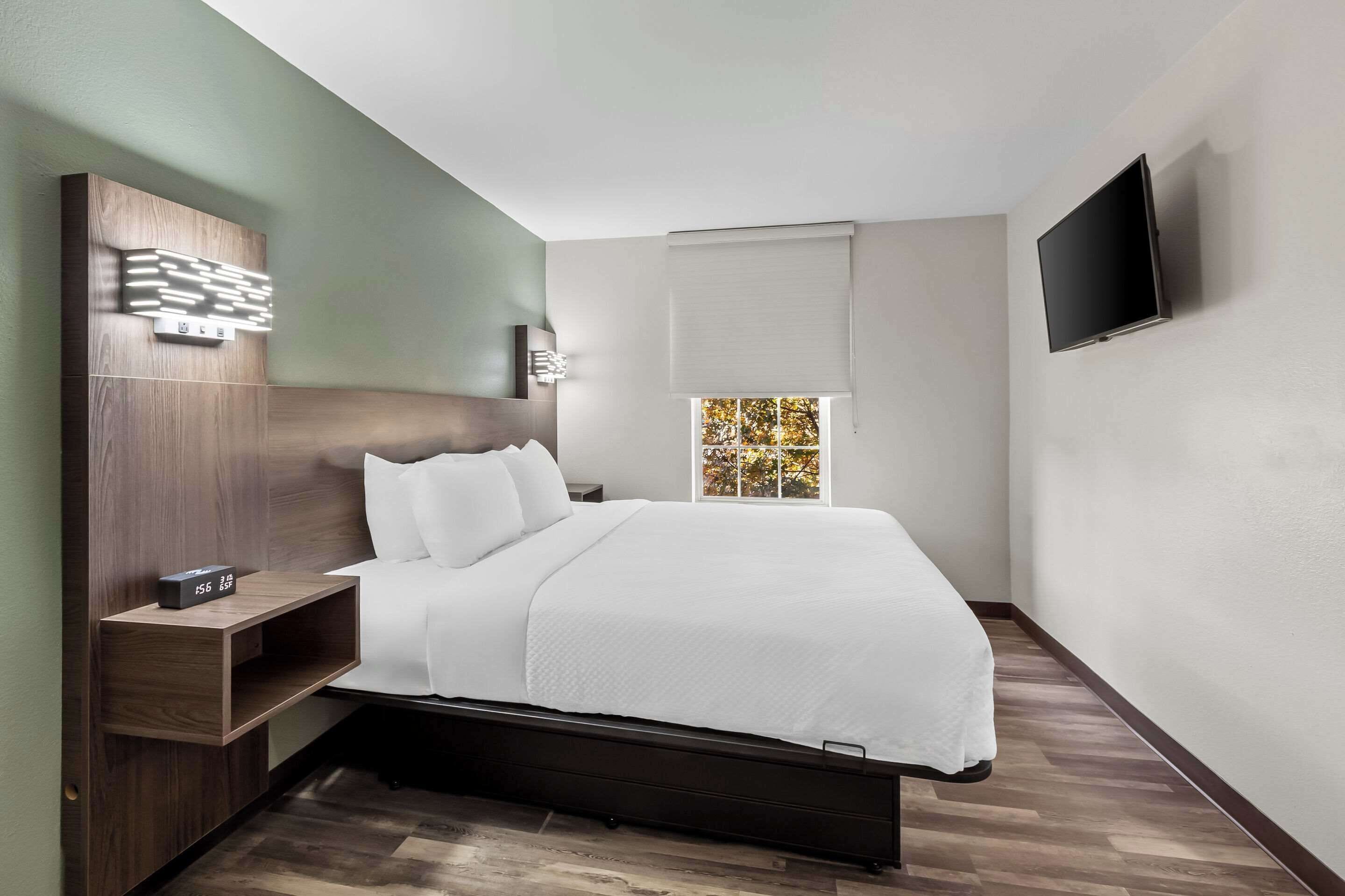 Una habitación de hotel moderna con un cabecero de madera, un televisor de pantalla plana montado en la pared, una mesita de noche con un reloj y una ventana con persianas.