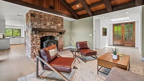 A modern living room with a stone fireplace, leather armchairs, a wooden coffee table, and a view of the kitchen and dining area.
