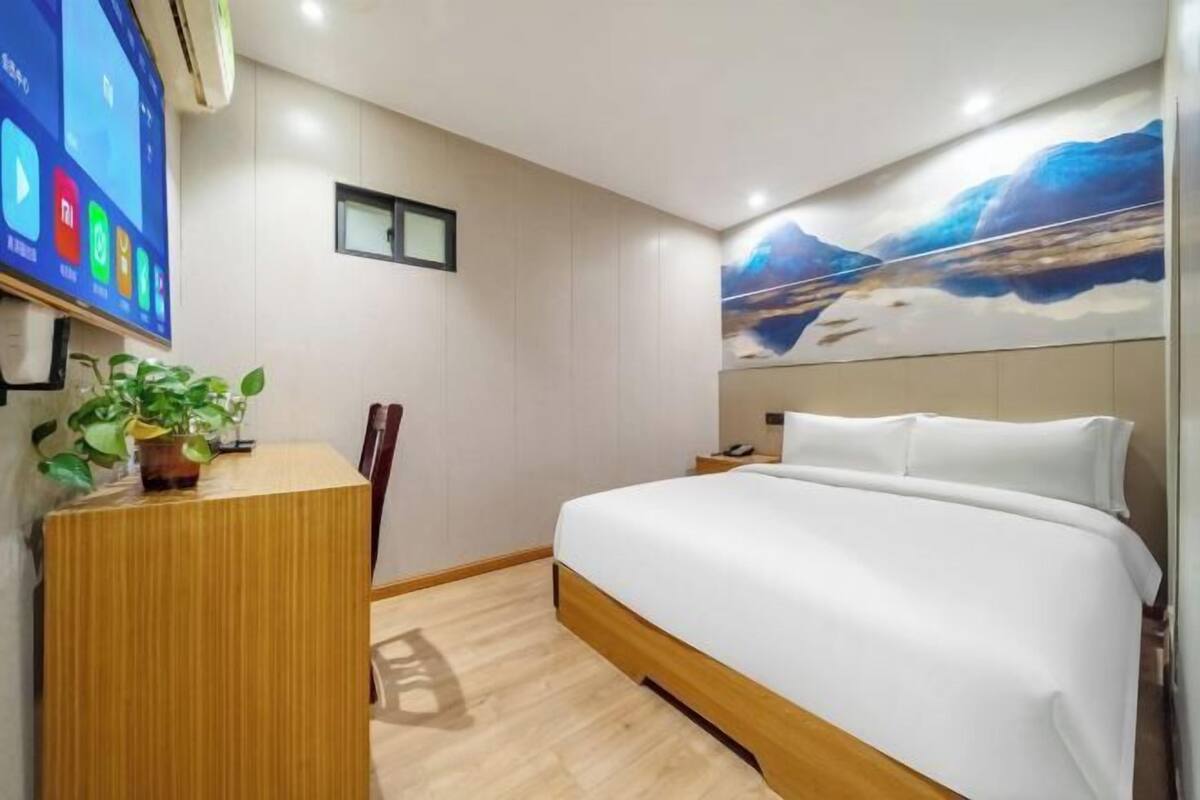 Quarto de hotel com cama, escrivaninha, cadeira, planta em vaso, TV na parede e pintura de montanhas.