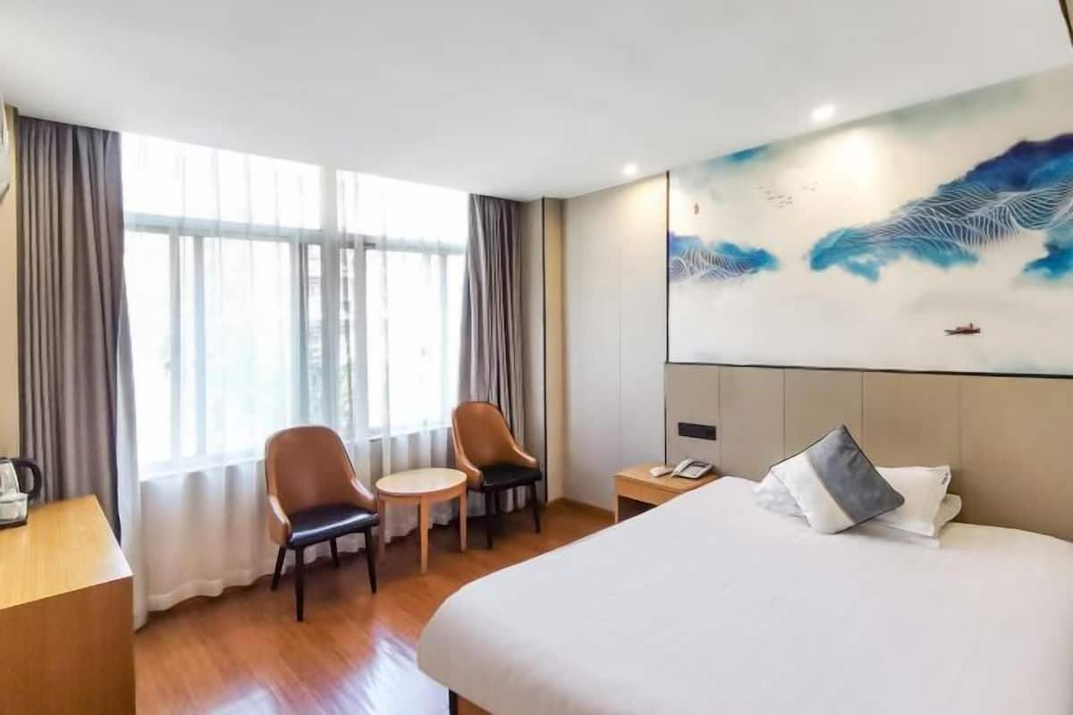 Quarto de hotel com uma cama grande, duas cadeiras, uma mesinha, uma mesa de cabeceira e um quadro na parede.