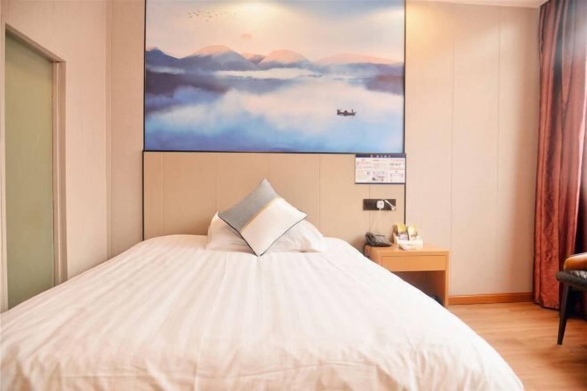 Uma cama bem arrumada com um travesseiro, uma mesa de cabeceira com abajur e um quadro de uma paisagem montanhosa na parede.