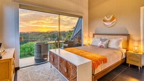 A bedroom with a large bed, a nightstand, a bedside table, and a view of a lush landscape through a sliding glass door.