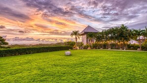 A lush green lawn with a stone centerpiece, a building with a pyramid roof, and a tropical garden with palm trees and string lights.