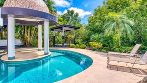 A swimming pool with a tiled deck, a covered seating area, and lush greenery in the background.