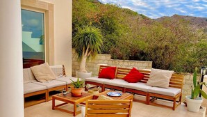 Un patio con sofá, sillas y mesa, rodeado de plantas y vistas a colinas.