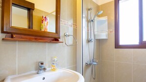 Un baño con lavamanos blanco, un espejo, ducha con cabezal de ducha manual y una ventana.