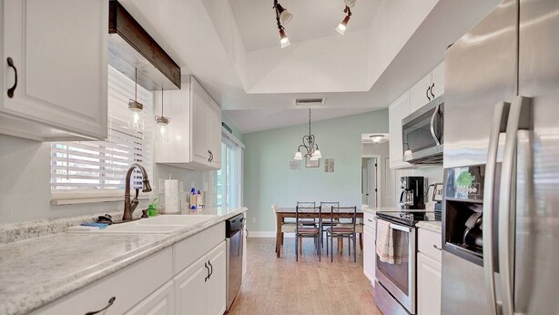 Una cocina moderna con armarios blancos, encimera de granito, electrodomésticos de acero inoxidable y una zona de comedor con mesa y sillas de madera.