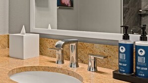 A bathroom with a granite countertop, a white sink, a mirror, a towel dispenser, and two bottles of Roots body lotion. - Grand Desert 1 Bedroom Suite, Las Vegas! (Las Vegas)