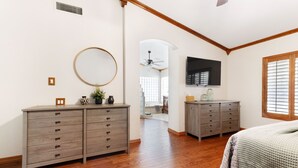 Un dormitorio con piso de madera, una cama con colcha, un aparador, un espejo, un televisor y una ventana con persianas.