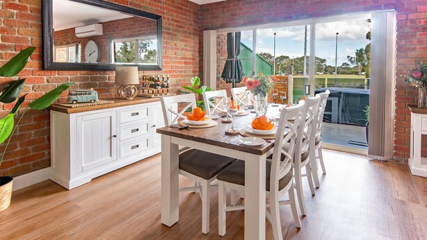 A dining area with a wooden table set for four, white chairs, a brick wall, a window, and a view of the outdoors.