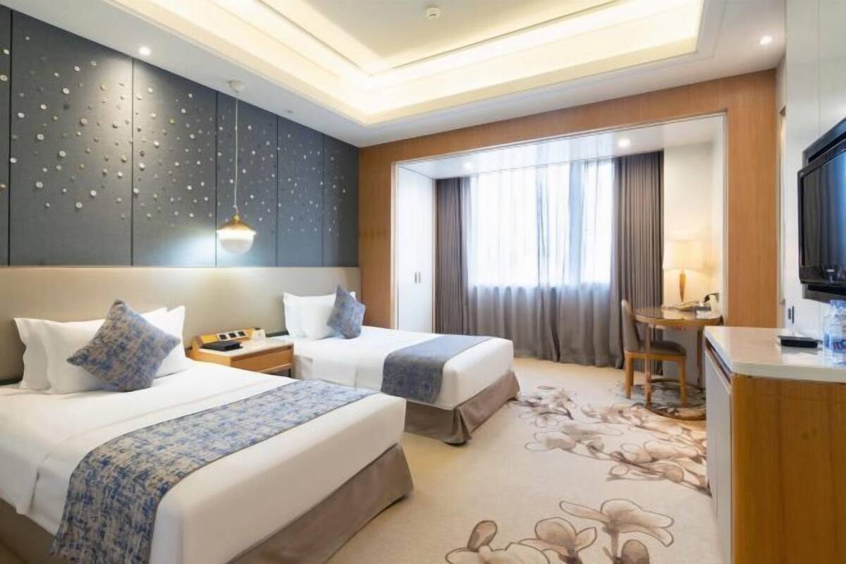 A hotel room with two beds, a desk, a TV, and a large window with curtains.