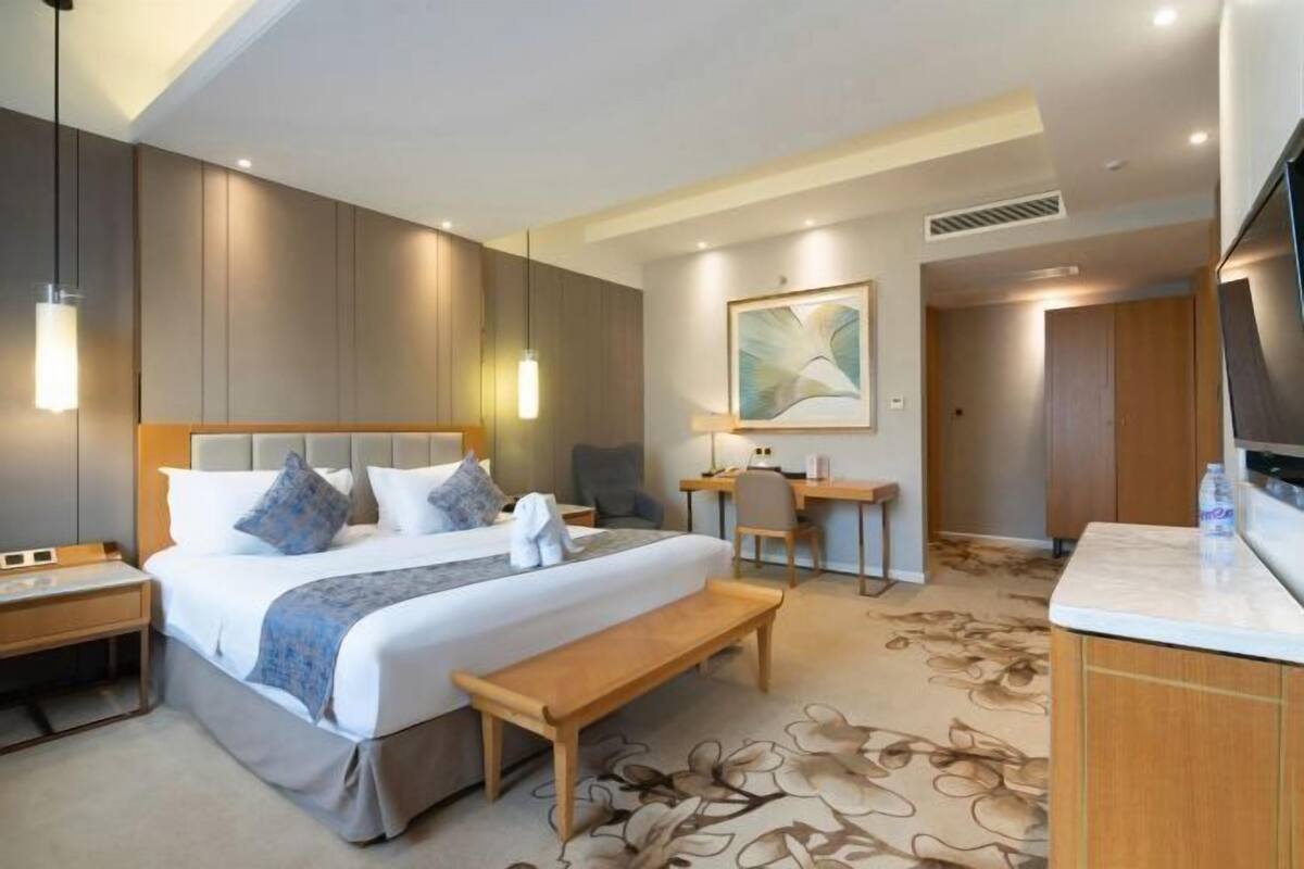 A hotel room with a large bed, bedside tables, a desk, a chair, a bench, a TV, and a painting on the wall.