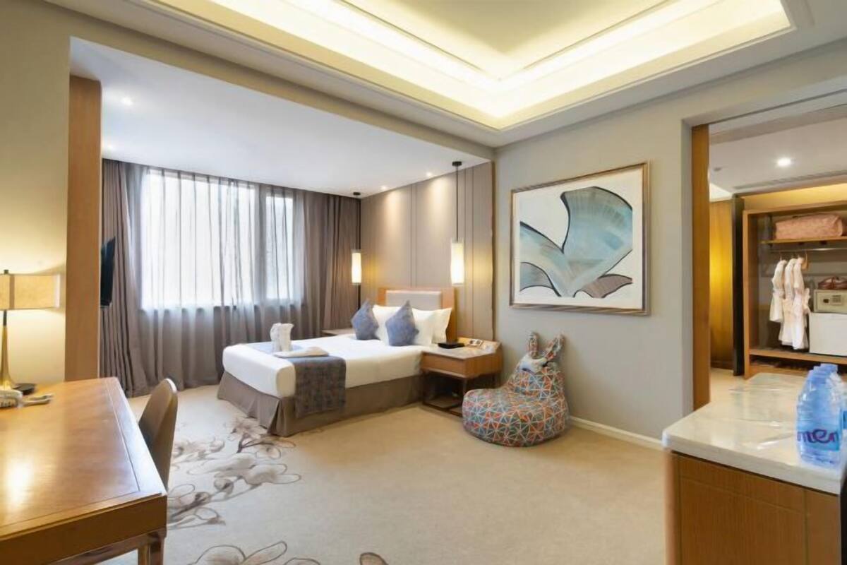 A hotel room with a bed, bedside table, lamp, chair, and a large abstract painting on the wall.