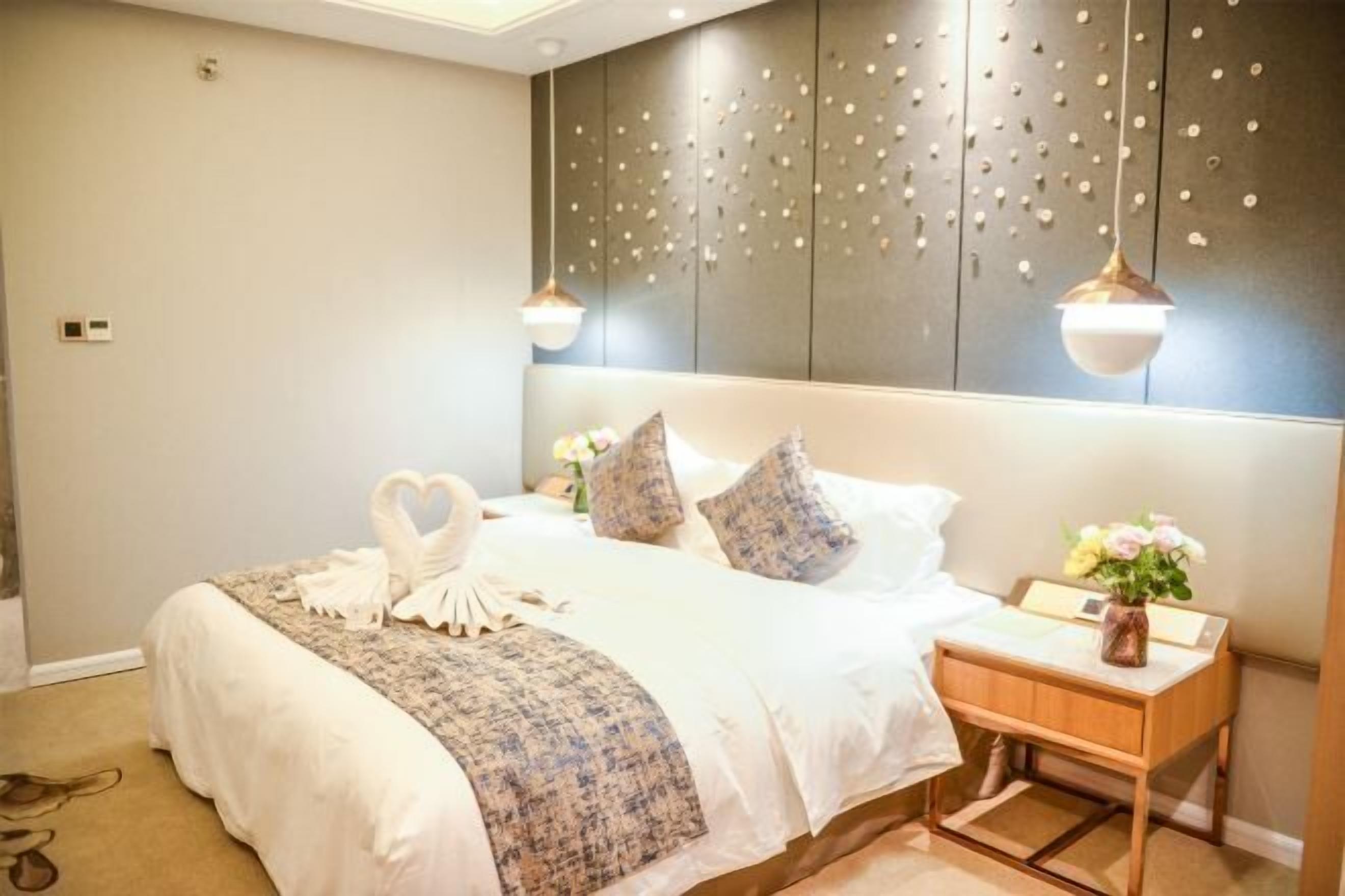 A neatly made bed with a heart-shaped towel arrangement, a bedside table with a vase of flowers, and a wall with a decorative panel.