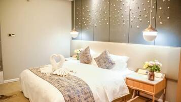 A neatly made bed with a heart-shaped towel arrangement, a bedside table with a vase of flowers, and a wall with a decorative panel.