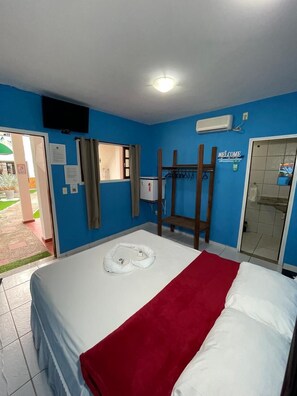 A hotel room with a bed, a red and white bedspread, a white towel, a wooden chair, a window with curtains, a wall-mounted air conditioner, and a 'Welcome' sign.