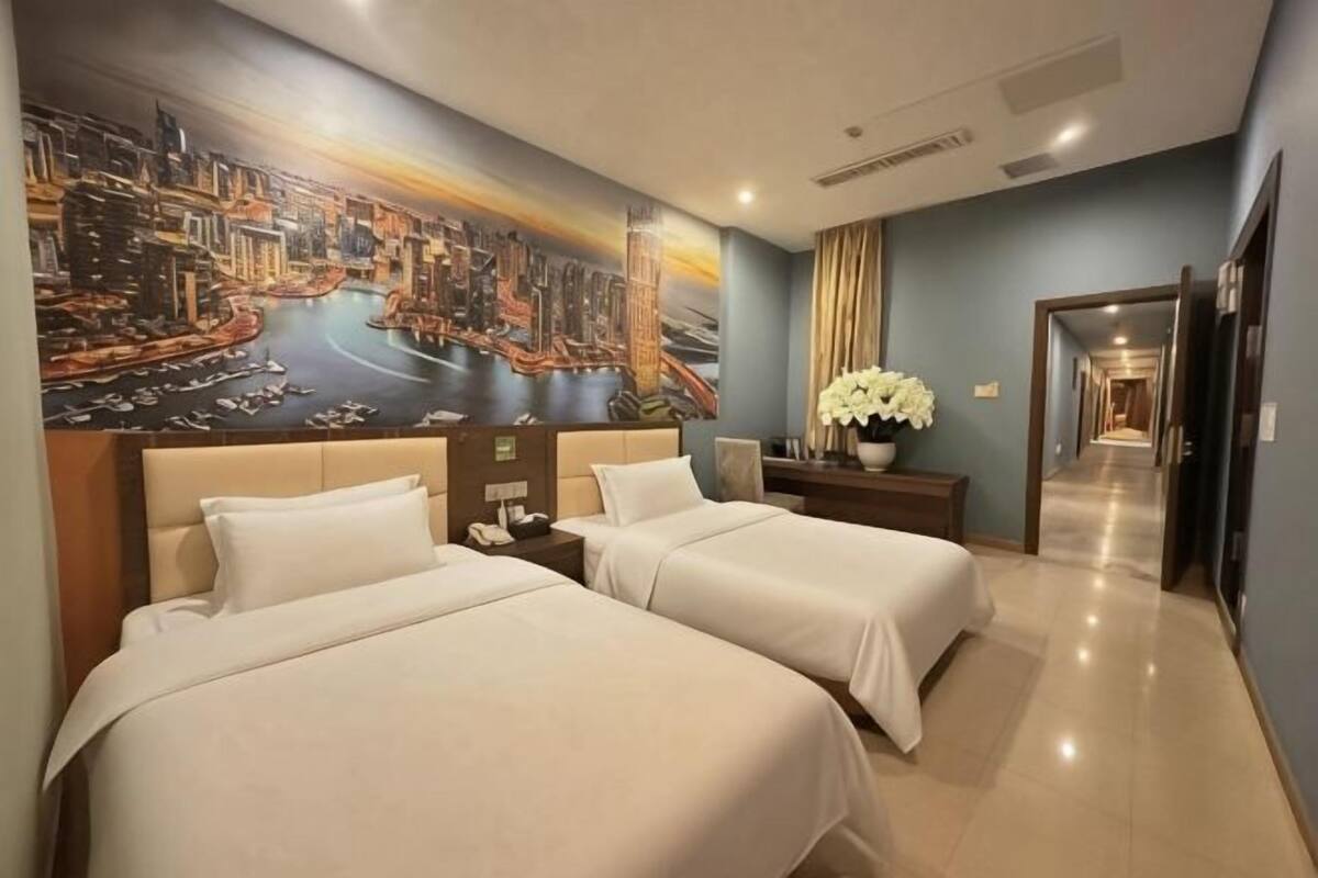 Une chambre d’hôtel avec deux lits, une fresque murale représentant un paysage urbain, une table de chevet et un couloir menant à la sortie.