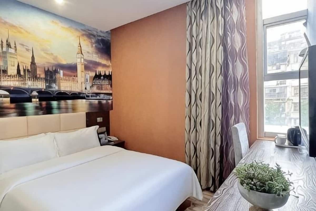 Une chambre d’hôtel avec un lit, un tableau représentant un paysage urbain, une table à manger avec un pot de plantes et une fenêtre avec des rideaux.