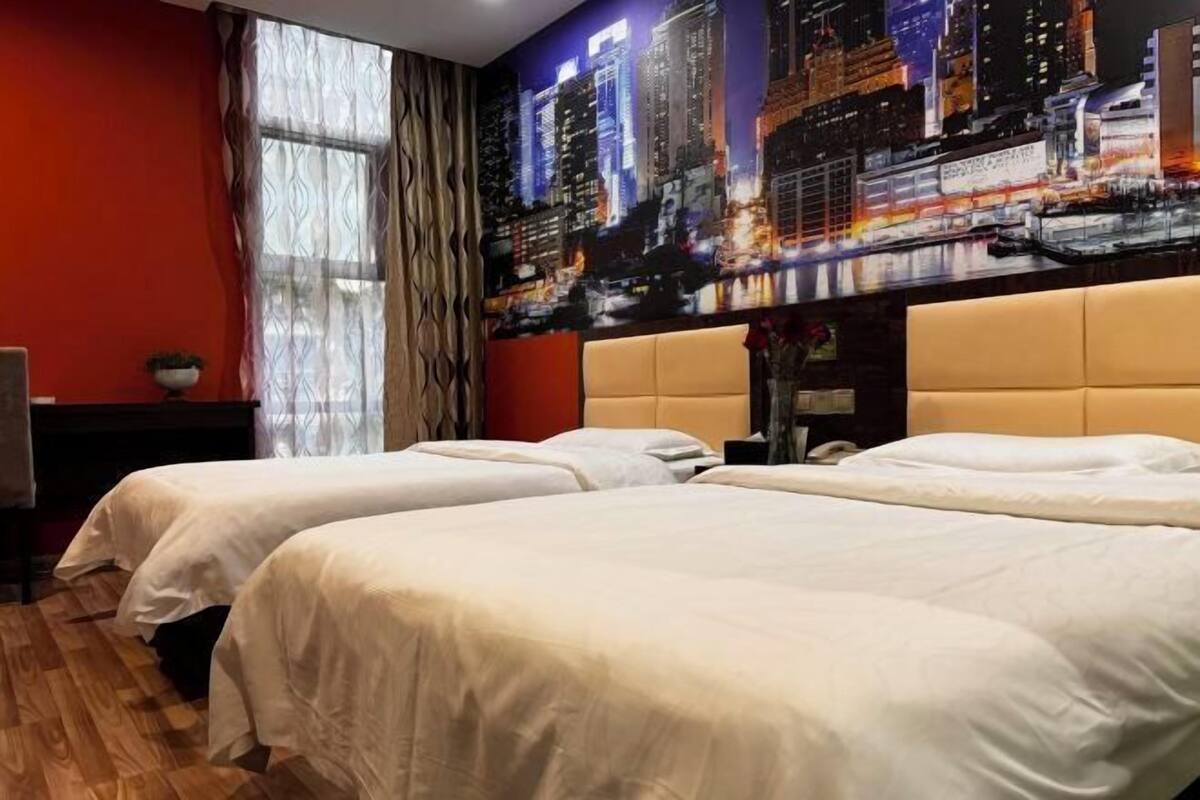 Une chambre d’hôtel avec deux lits, une fresque représentant un paysage urbain sur le mur, une fenêtre avec des rideaux et un plancher en bois.