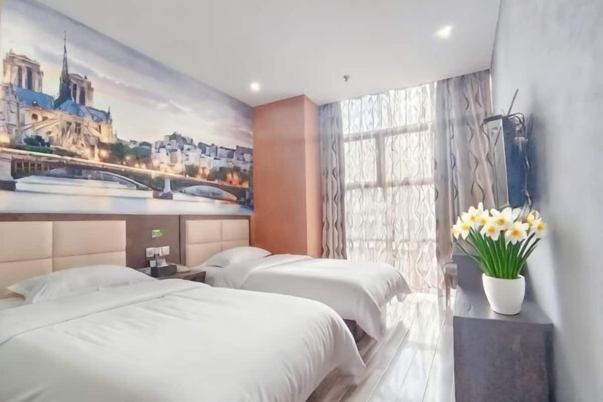 Une chambre d’hôtel avec deux lits, une fresque représentant un paysage urbain, un vase de fleurs et une fenêtre avec des rideaux.