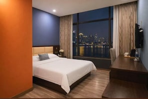 A hotel room with a bed, a desk, a view of a cityscape at night, and a large window.