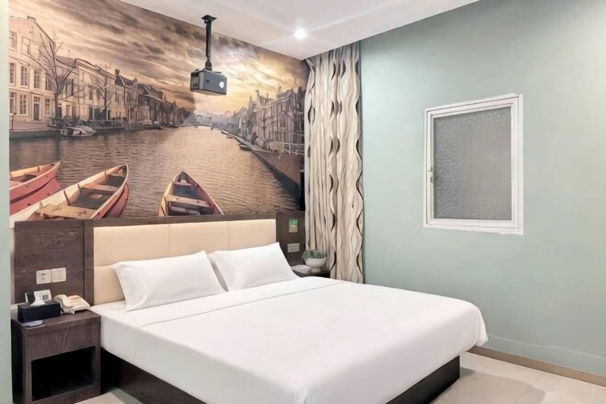 Une chambre d’hôtel avec un lit, une table de chevet, un projecteur fixé au mur et une fenêtre avec des rideaux.